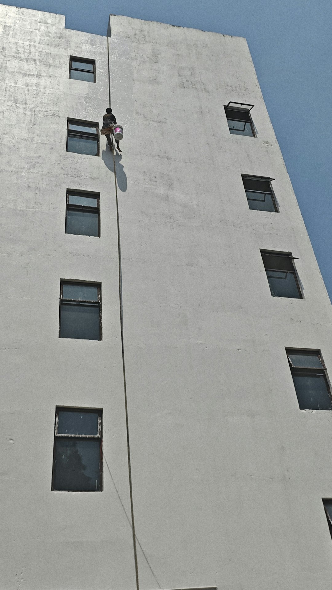 A high-angle, vertical shot of a tall, white building facade under a clear blue sky. A lone worker is suspended mid-air by a single rope, positioned between rows of dark, rectangular windows. Carrying a bucket, the figure appears small against the vast, weathered expanse of the concrete wall, highlighting the grueling nature of high-rise maintenance. The composition emphasizes clean lines, repetitive geometric patterns, and the dramatic contrast between the bright architectural surface and the dark window frames.